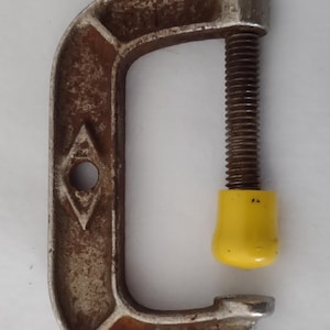 May include: A C-clamp with a rusty, metallic frame and a threaded rod. The clamp has a yellow protective cap on the end of the rod. The tool is designed to hold objects together.