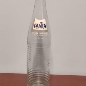 May include: Clear glass Fanta bottle with a white and blue label. The label features the Fanta logo and the text "RETURN FOR DEPOSIT". The bottle has a ribbed design and is 10 fl oz.