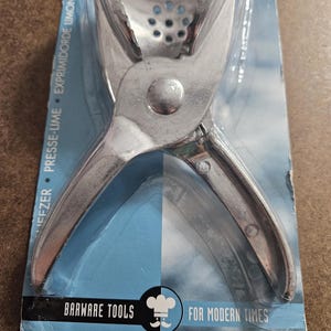 May include: A silver-colored citrus juicer in its original packaging. The juicer is made of metal and has a hinged design with a perforated bowl. The packaging is blue and white with the text "BARWARE TOOLS FOR MODERN TIMES".