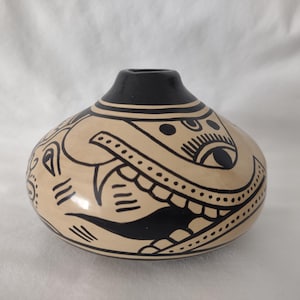 May include: A hand-painted ceramic vase with a rounded body and a narrow neck. The vase features a cream-colored base with black tribal-style designs, including stylized eyes and geometric patterns. The rim and neck are black.