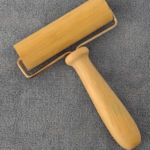 May include: A wooden brayer featuring a cylindrical roller attached to a sturdy handle by a metal frame. The tool has a light tan wood grain finish on both the roller and the handle, with a smooth, polished texture and a simple, functional design.