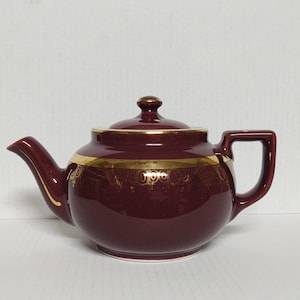 May include: A maroon teapot with a gold trim and a gold design around the middle. The teapot has a lid and a handle.