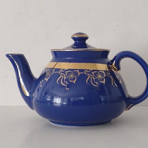 May include: A cobalt blue teapot with a gold band and floral design. The teapot has a curved spout, a rounded body, and a loop handle. The lid is topped with a small gold knob. The teapot is made of ceramic.
