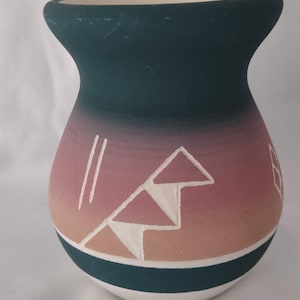 May include: A hand-painted ceramic vase with a wide base and a narrow neck. The vase features a gradient of colors, transitioning from a dark teal at the top to pink and tan in the middle. White geometric designs adorn the surface.