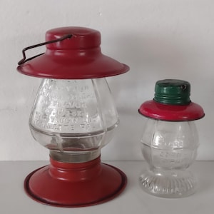 May include: Two vintage glass syrup dispensers with red metal lids and bases. The larger dispenser has a handle and text on the glass. The smaller dispenser has a green top. Both are on a white surface.