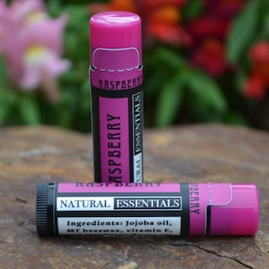 Könnte beinhalten: Zwei rosa Lippenbalsam-Tuben mit schwarzen Etiketten. Das Etikett auf der oberen Tube lautet "Raspberry Natural Essentials". Das Etikett auf der unteren Tube lautet "Raspberry Natural Essentials Zutaten: Jojobaöl, MT Bienenwachs, Vitamin E."