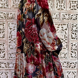 May include: A long, flowing duster cardigan featuring a dark black base with a vibrant, multi-colored floral print in shades of deep red, burgundy, and cream. The garment has long sleeves and is adorned with subtle, shimmering gold-toned beadwork.