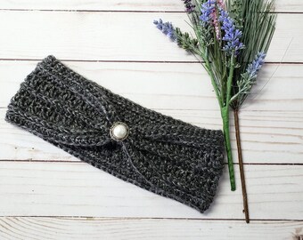 May include: A gray crocheted headband with a pearl button accent. The headband is lying on a white wooden surface.