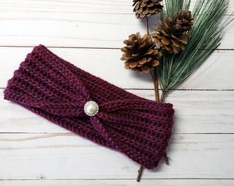 May include: A purple knitted headband with a pearl button accent. The headband is on a white wooden background with pine branches and pine cones.