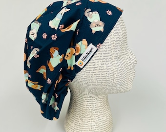 May include: A navy blue scrub cap with a repeating pattern of cartoon dogs, cats, and paw prints in tan, light blue, and pink. The cap has a tag that says "KimKaps".