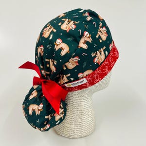 May include: A dark green scrub cap with a Christmas sloth pattern, featuring sloths wearing Santa hats and scarves, candy canes, and sprigs of holly. The cap has a red ribbon and a red patterned trim. The brand name "KimKaps" is visible.