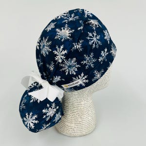 May include: A navy blue scrub cap featuring a snowflake pattern in white and silver. The cap has a white bow and the brand name "KimKaps" is visible. This cap is designed for medical professionals or those in the healthcare field.