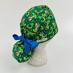May include: Green surgical scrub hat featuring a pattern of cats wearing leprechaun hats and holding pots of gold at the end of rainbows. The hat has a blue ponytail holder and a label that reads "KimKaps".