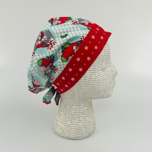 May include: A festive scrub cap with a light blue pattern of cats in stockings and white polka dots. The cap has a red band with white snowflakes. It is designed to tie at the back.