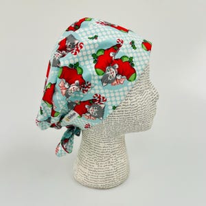 May include: A blue and white patterned surgical scrub hat with a Christmas theme. The hat features a repeating pattern of kittens wearing Santa hats and candy canes. The hat is tied with a blue ribbon.