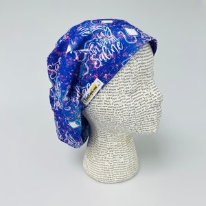 May include: A blue and white scrub cap with the text "Give Normal Saline" and medical-themed graphics. The cap has a galaxy-like pattern with pink and white accents. The brand name "KimKaps.com" is visible on a small tag.