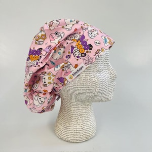 May include: A pink scrub cap featuring a repeating pattern of cartoon ghosts, skulls, and spiderwebs with the text "Boo Crew" in purple and orange. The fabric head covering includes a white tag with the brand name KimKaps printed in black.