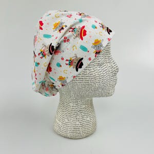 May include: White scrub hat with a colourful print of cartoon cats wearing cowboy hats and boots. The print includes hearts, horseshoes, and cacti.