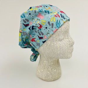 May include: A blue and white patterned surgical scrub hat with a tie. The pattern features various summer-themed items like swimsuits, flip-flops, and palm trees.