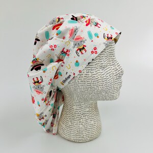 May include: A white scrub cap with a repeating pattern of cartoon cowboys, cacti, and flowers. The cap has a tie-back design and the text "KimKaps". The cap is displayed on a mannequin head.