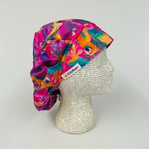May include: A scrub cap featuring a vibrant tropical pattern with bright pink, orange, and green colors, showcasing parrots and lush foliage. The fabric head covering includes a white rectangular label with the text KimKaps printed in black.