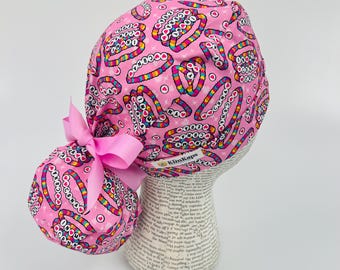 May include: A pink scrub cap with a repeating pattern of colorful bracelets and circles. The cap has a matching pink bow and is displayed on a mannequin head. The brand name "KimKaps" is visible on a tag.