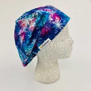 May include: A blue, purple, and white patterned surgical scrub cap with a snowflake design. The cap is made of fabric and has a tag that reads "KimKaps". The cap is displayed on a mannequin head.