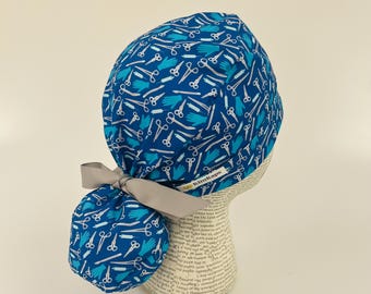 Op de afbeelding: Een blauwe chirurgische scrubcap met een patroon van witte scharen en blauwe handschoenen. De cap heeft een grijs lint en de merknaam "KimKaps" is zichtbaar. De cap wordt getoond op een mannequinhoofd.