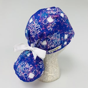 May include: A blue scrub cap with a white ribbon and the words "Salty Live Normal Saline" in white. The cap has a pattern of white saline bags and text on a blue and pink background.