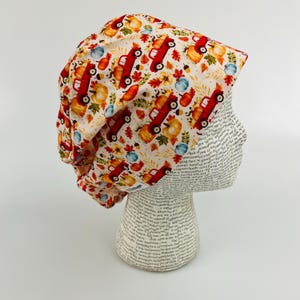 May include: A white and cream-colored scrub cap with a repeating pattern of red trucks, pumpkins, and fall foliage. The cap is made of a soft fabric and is designed to be worn in a medical or professional setting.