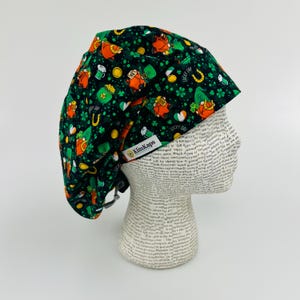 Peut inclure: Un bonnet de travail noir avec un imprimé vert, blanc et orange de lutins, trèfles, pièces d'or et le texte "Lucky Day". Le bonnet a un cordon de serrage et une étiquette qui dit "KimKaps".