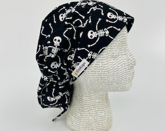 Op de afbeelding: Zwarte chirurgische scrubcap met een herhaald patroon van witte skeletten. De cap heeft een gebogen rand en een label met de tekst "KimKaps". Ontworpen om het haar te bedekken tijdens medische procedures.