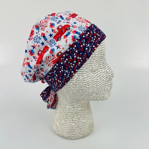 May include: A red, white, and blue surgical scrub hat with a red truck, fireworks, and stars pattern. The hat has a blue and white star pattern on the back.