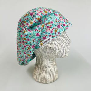 May include: A floral print scrub cap in shades of teal, pink, yellow, and orange. The cap is displayed on a mannequin head. The tag on the cap reads "KimKaps".