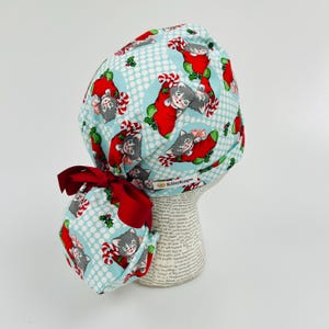 May include: A blue and white patterned surgical scrub hat with a red bow and a red and white candy cane design. The hat features a repeating pattern of kittens wearing Santa hats and holding candy canes. The hat has a label that says "KimKaps".