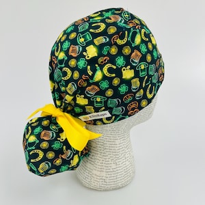 May include: A dark green scrub cap with a repeating pattern of beer mugs, horseshoes, and shamrocks in neon yellow and orange. A yellow bow is attached to the back. The brand name "KimKaps" is visible on a small tag.