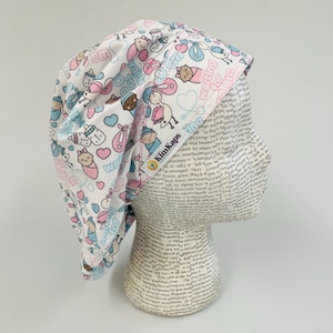 May include: A white surgical scrub cap with a baby-themed pattern. The cap features illustrations of babies, bottles, and hearts in pink, blue, and brown. The words "waiting for" are printed on the cap.