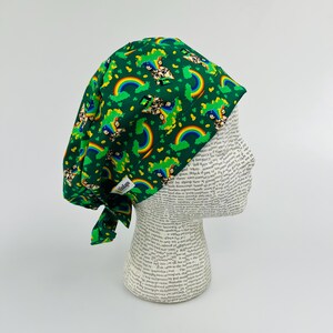 May include: Green scrub cap with a St. Patrick's Day theme. The cap features a pattern of rainbows, leprechauns, and shamrocks. The cap is tied at the back and has a small tag that says "TimKaps".