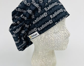 Op de afbeelding: Een zwarte scrub-muts met het woord "Radiologie" herhaald in witte en grijze tekst. De muts heeft een zwart elastiek en een wit label met de tekst "KimKaps".