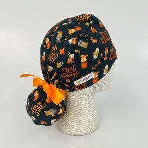 May include: A black scrub cap featuring a repeating pattern of orange and white autumn-themed coffee cups, leaves, and the text "Happy Fall" in orange script. The cap includes a bright orange ribbon tie and a white label reading "KimKaps".
