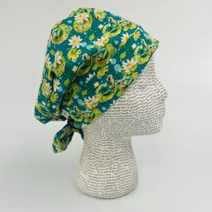 May include: A teal scrub cap featuring a pattern of lily pads, white water lilies, and green frogs. The cap is tied at the back and is displayed on a mannequin head.