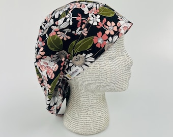 Op de afbeelding: Een zwarte scrubmuts met een bloemenprint met roze, witte en groene bloemen. De muts heeft een wit elastiek en de tekst "ScrubKaps" is aan de zijkant gedrukt.