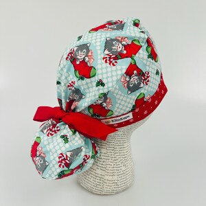 May include: A light blue surgical scrub cap with a red bow and trim. The cap features a repeating pattern of gray kittens in Christmas stockings, candy canes, and holly on a polka dot background. The brand name "KimKaps" is visible.