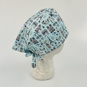 May include: A light blue surgical scrub hat with a Parisian themed print. The print features the Eiffel Tower, bicycles, baguettes, and other Parisian icons. The hat has a tie at the back.