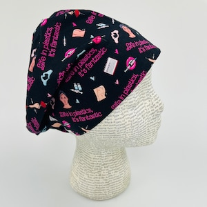 May include: Black surgical scrub cap with a repeating pattern of cosmetic surgery-related illustrations in pink and white. The cap features the phrase "Life in plastics, it's fantastic" in pink lettering.