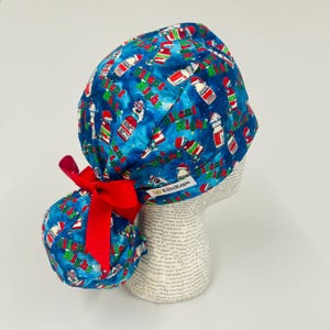 May include: A blue scrub cap with a Christmas-themed pattern featuring candy canes, snow, and the words "Silent Night." The cap has a red bow and is displayed on a mannequin head.
