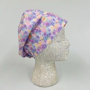 May include: A floral patterned scrub cap in shades of purple, pink, and yellow. The cap is made of fabric and features a watercolor-like design of roses and other flowers. The cap is displayed on a mannequin head.