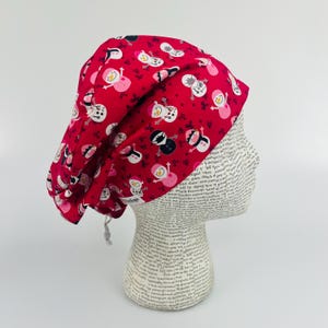 May include: Red scrub hat with a snowman pattern. The hat has a drawstring tie.