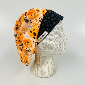 May include: A scrub cap featuring a light orange fabric with a pattern of jack-o'-lanterns, white flowers, and black bats. The cap has a solid black band with a repeating heart pattern and a white fabric tag that reads KimKaps.