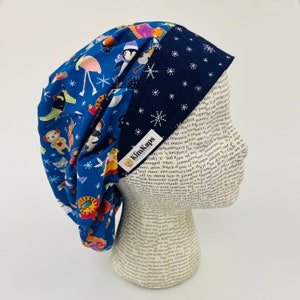 May include: A reversible blue and navy scrub cap. One side features a pattern of cartoon animals and snowflakes on a blue background. The other side is navy blue with white snowflakes. The label reads "KimKaps".